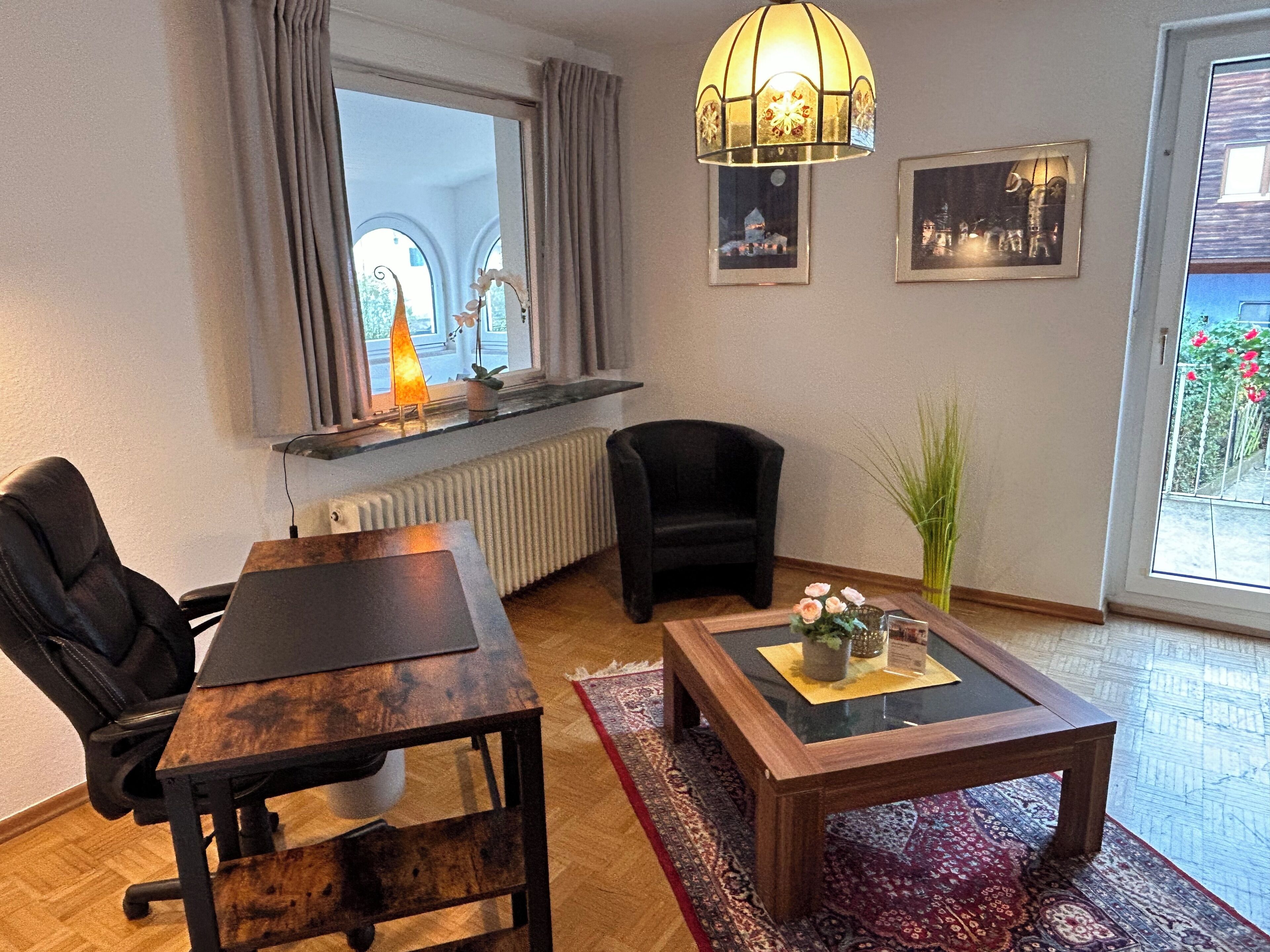 Una sala de estar acogedora con una mesa de madera, un sillón negro, una mesa de centro de vidrio, un radiador, una ventana con cortinas y una lámpara decorativa.