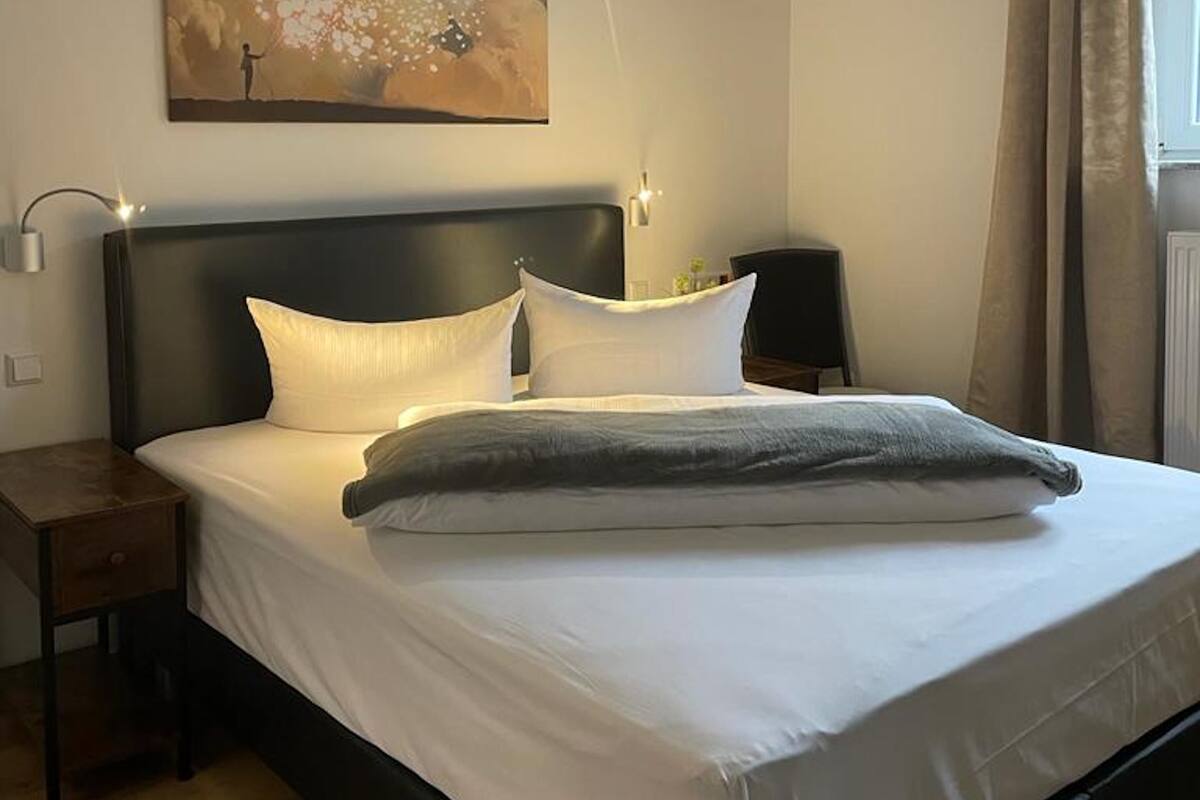 Ein ordentlich bezogenes Bett mit weißen Leinen und einer grauen Tagesdecke, ein Nachttisch mit Lampe sowie ein Gemälde an der Wand.