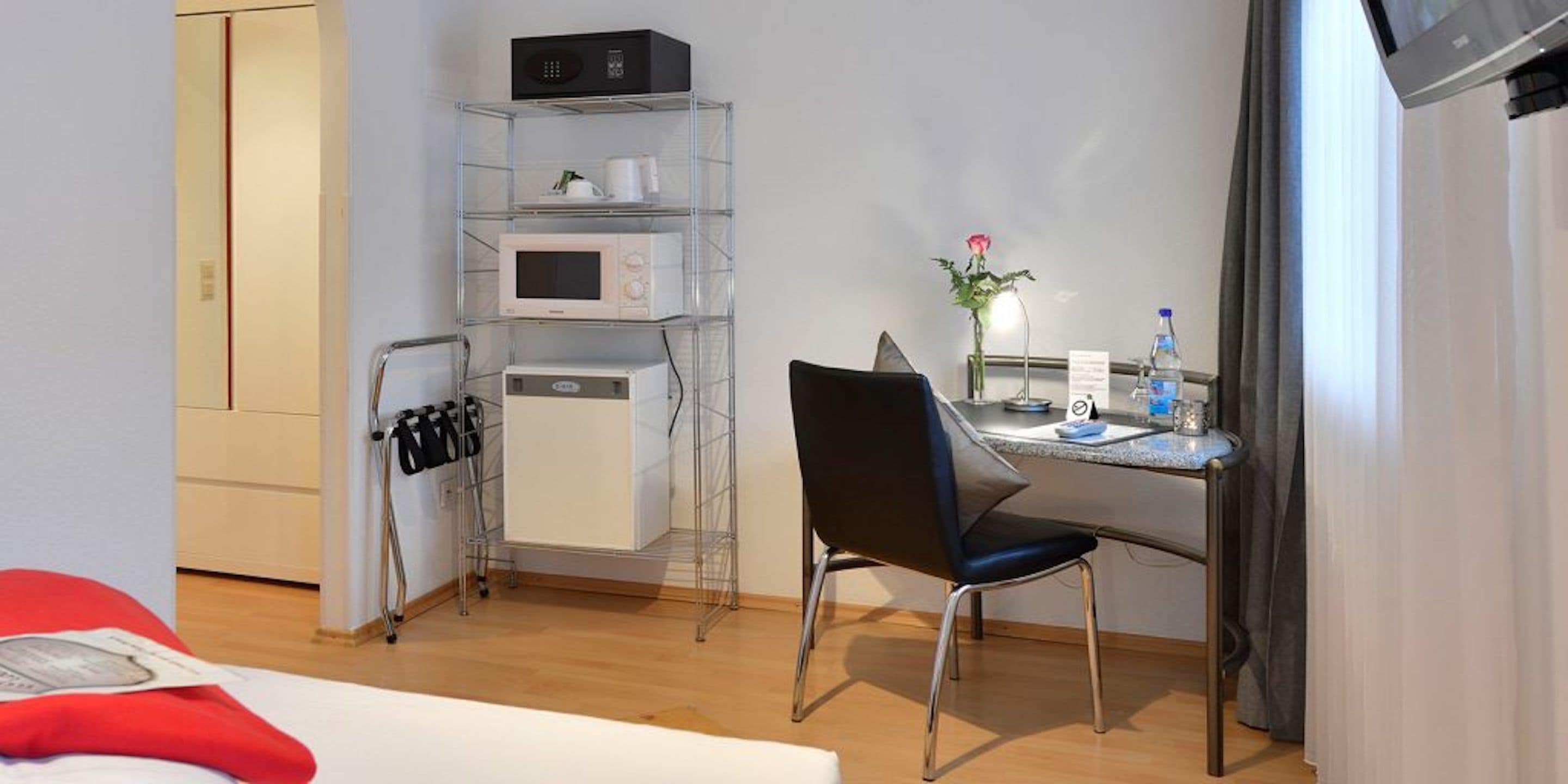 Habitación de hotel con cama, escritorio con silla, microondas, cafetera, botella de agua y un florero con una flor.