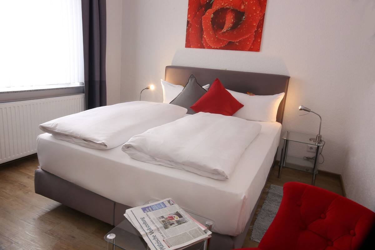 Ein Doppelbett mit weißen Bettwäsche, einem roten Kopfkissen und einer Zeitung auf einem Glastisch.