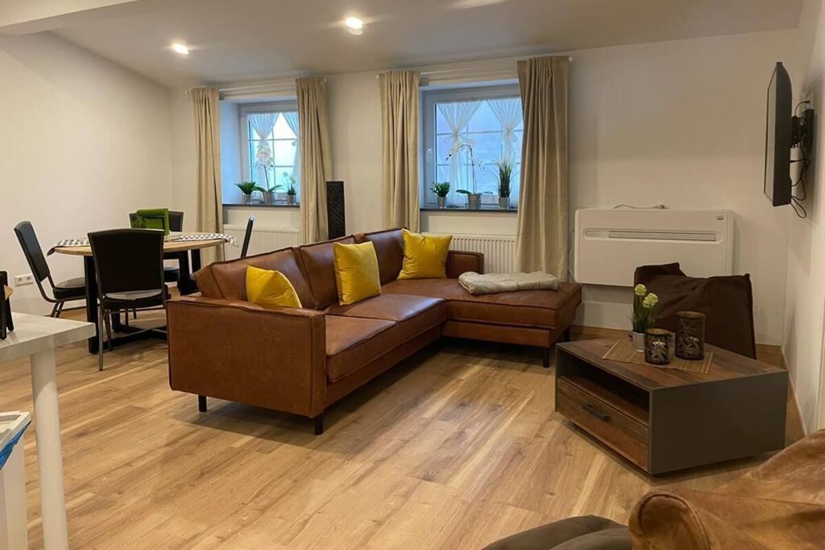 Ein modernes Wohnzimmer mit einem braunen Ecksofa, einem Couchtisch aus Holz und einem Essbereich mit Tisch und Stühlen.