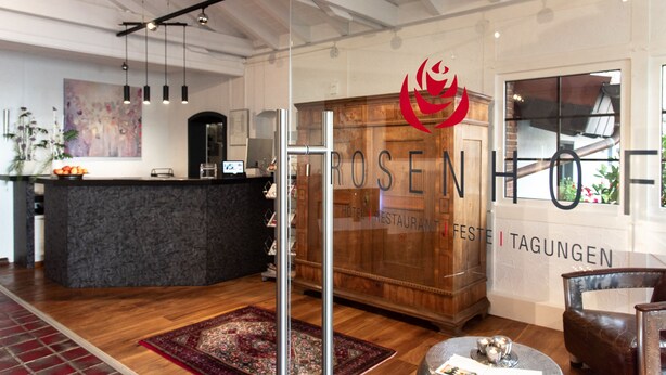 Un hall d’hôtel moderne avec un comptoir d’accueil, un espace de réception en bois, une grande fenêtre et un logo rouge sur le mur.