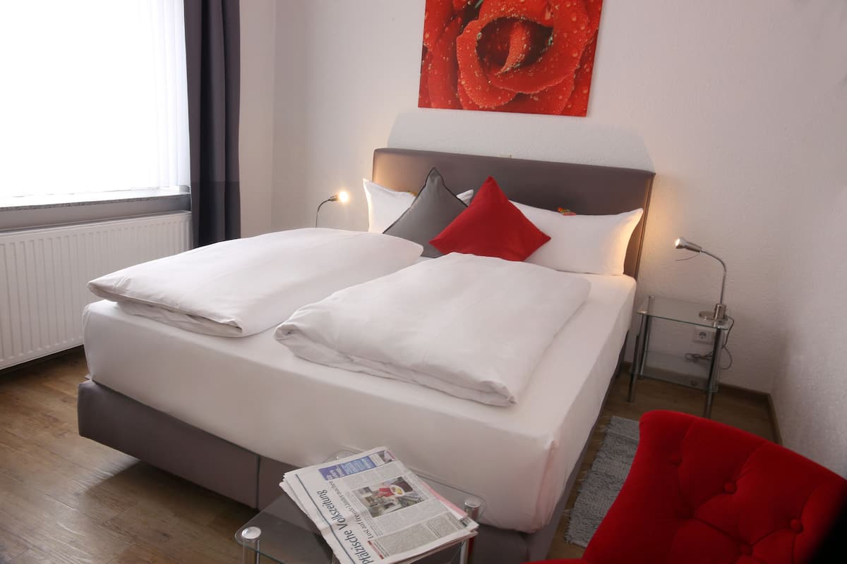Ein Doppelbett mit weißer Bettwäsche, einem roten Kissen und einem grauen Kissen. Ein Glastisch mit einer Zeitung und ein roter Stuhl.