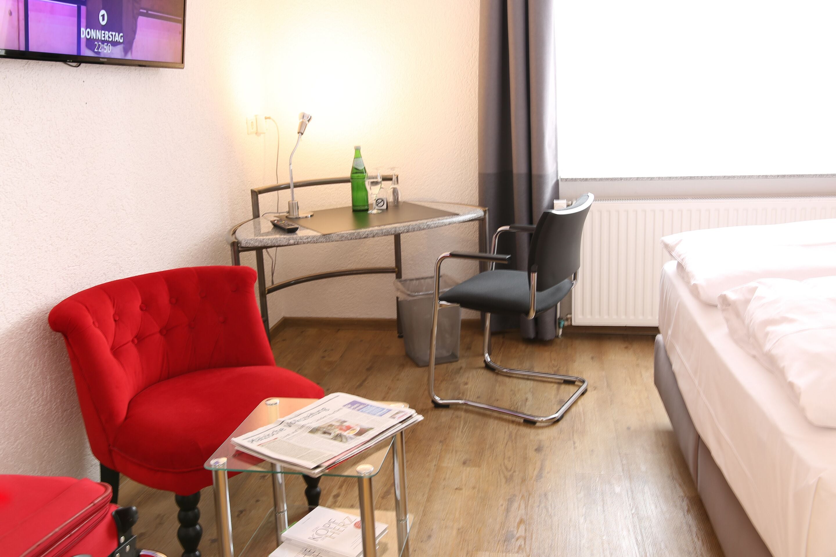 A hotel room with a bed, a red armchair, a small table with a newspaper, a chair, and a TV on the wall.