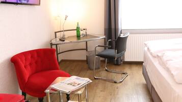 A hotel room with a bed, a red armchair, a small table with a newspaper, a chair, and a TV on the wall.