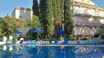 Zona de piscina con sillas de descanso y sombrillas, rodeada de altos árboles y un edificio de varios pisos con balcones.