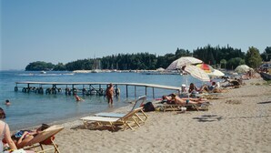 A beach with people sunbathing and swimming, a pier extending into the water, and a row of beach umbrellas. - Ipsos Beach (Corfu)