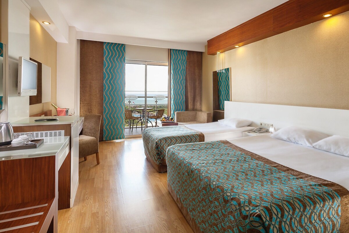 A hotel room with two beds, a desk with a telephone, a chair, and a view of the sea.