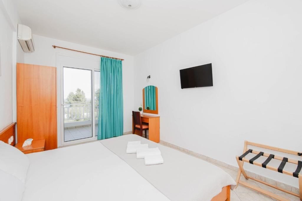 Habitación de hotel con cama, escritorio, silla, televisor y balcón con cortinas.