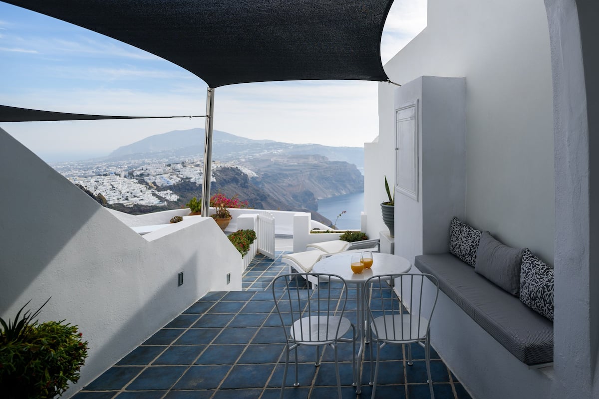 A balcony with a white wall, blue tiles, a table with two chairs, a sofa with patterned cushions, and a view of a coastal landscape.