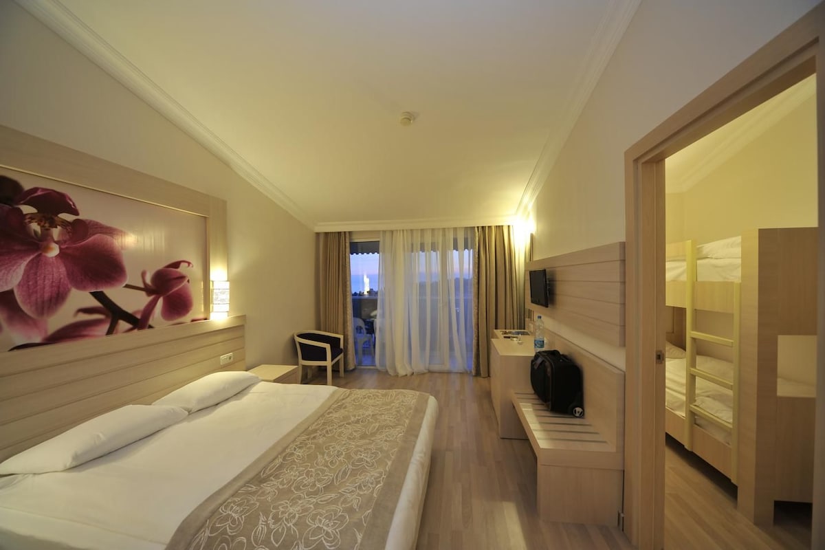 A hotel room with a large bed, a desk, a chair, a TV, and a view of the city through the window.
