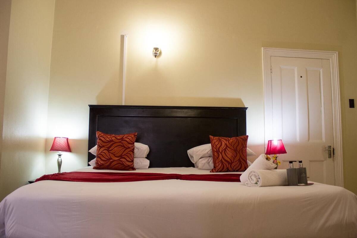 Un lit bien fait, avec une tête de lit sombre, deux coussins à motifs et un plaid rouge. On y trouve deux lampes de chevet et une serviette pliée.