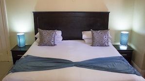 Un lit bien fait, avec une tête de lit sombre, deux coussins à motifs et deux lampes de chevet dotées d’abat-jour à motifs bleus.