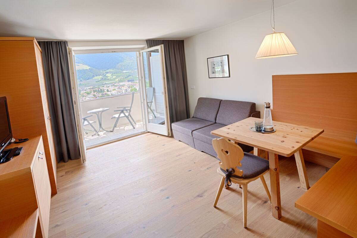 Habitación de hotel con piso de madera, un sofá, una mesa de comedor con una silla, un televisor y un balcón con vista al paisaje.