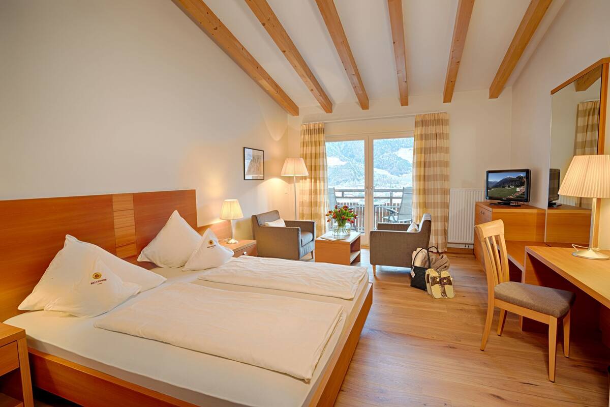 Habitación de hotel con techo de madera, cama con ropa blanca, escritorio con silla, televisión y vista a las montañas a través de las ventanas.
