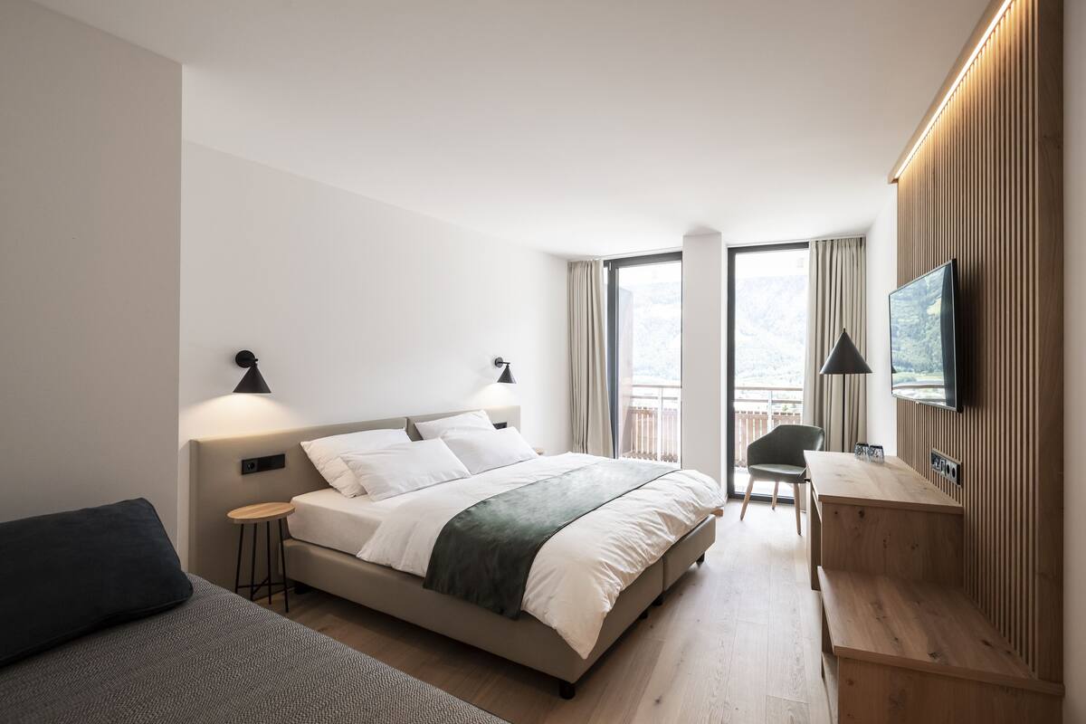 Una habitación de hotel moderna con cama, lámparas de noche, televisión, una mesita y una silla.