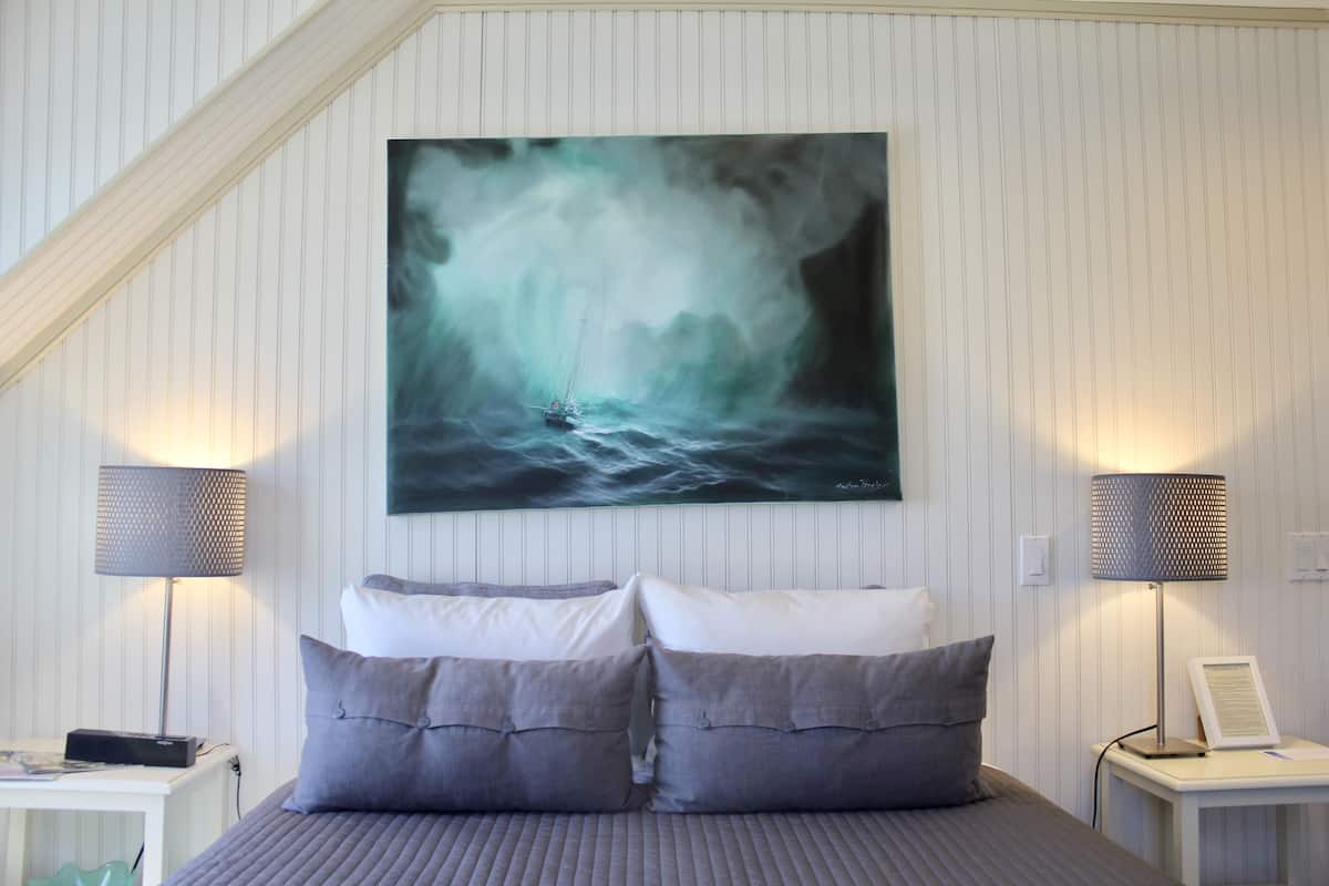 A bedroom with a bed, two bedside lamps, a nightstand, and a wall-mounted painting of a ship in a stormy sea.
