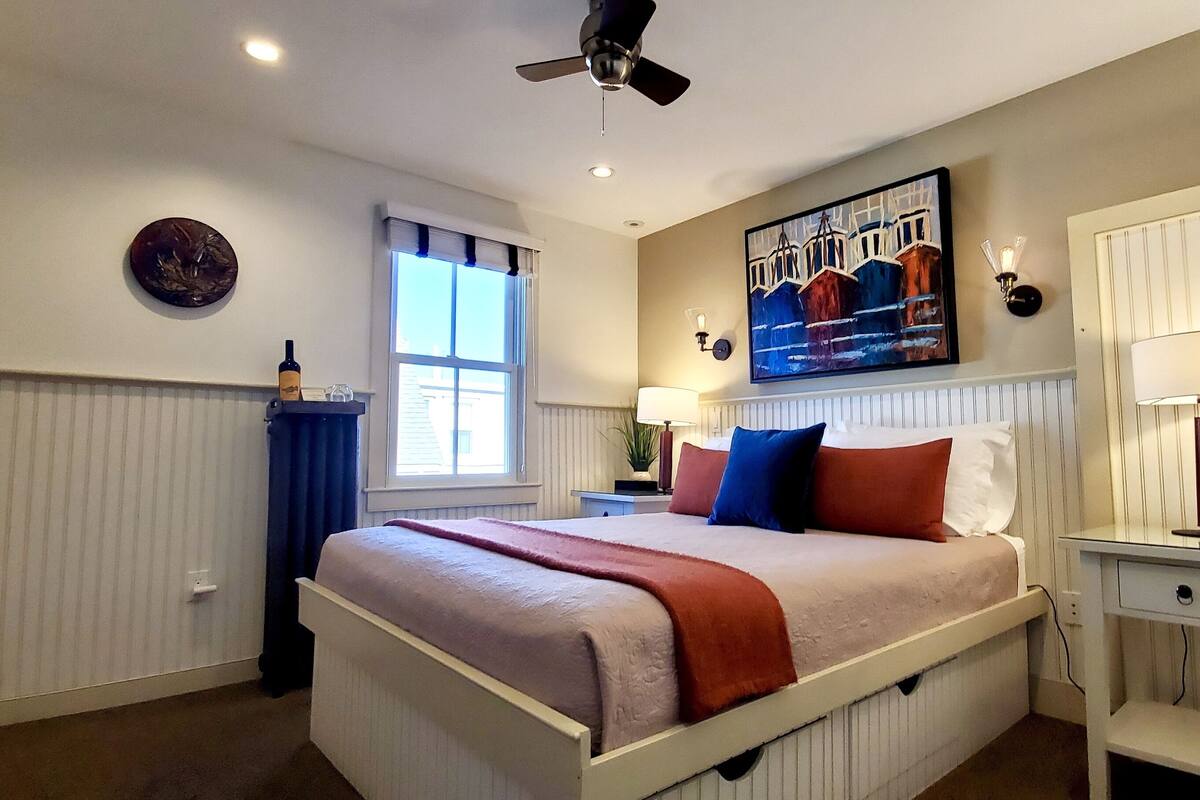 A bedroom with a bed, a window with curtains, a ceiling fan, a painting on the wall, and a bedside table with a lamp.
