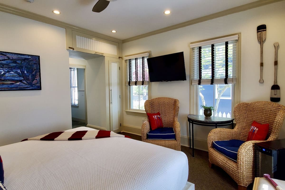 A hotel room with a large bed, two wicker chairs, a small table, a TV, and a painting on the wall.