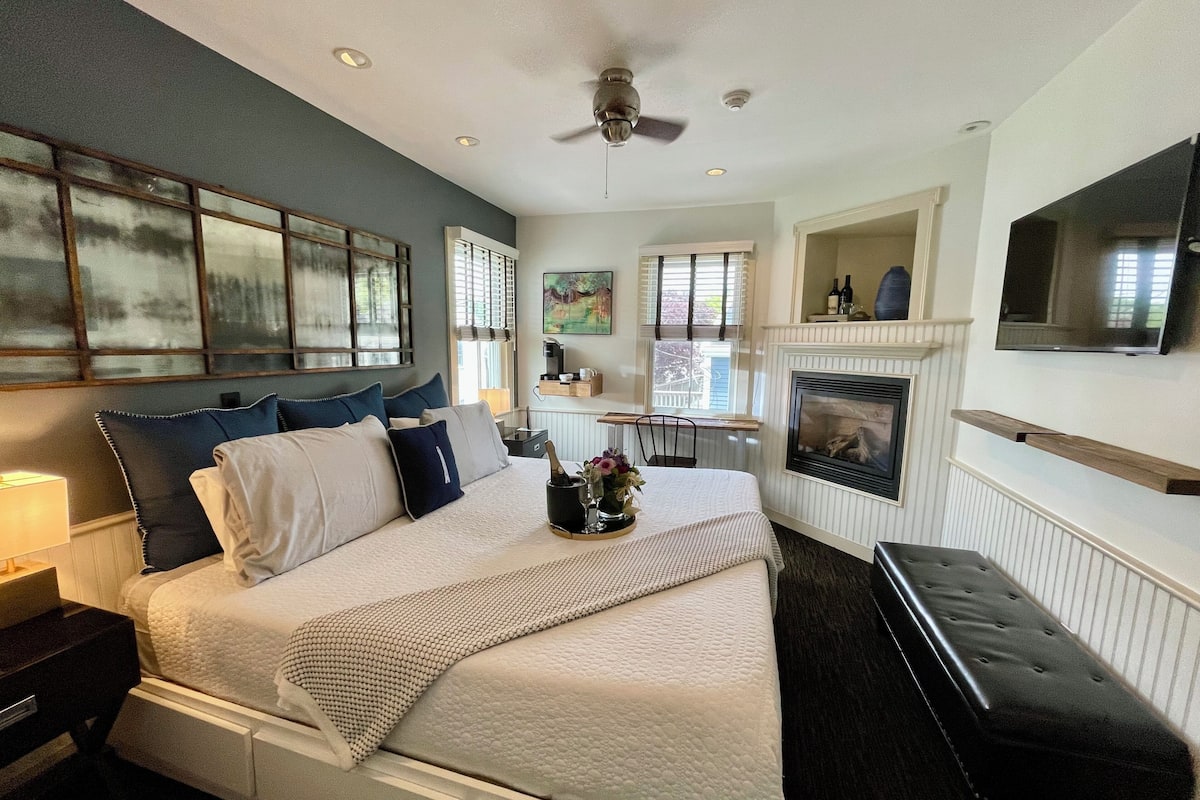 A bedroom with a large bed, a ceiling fan, a fireplace, and a TV mounted on the wall.