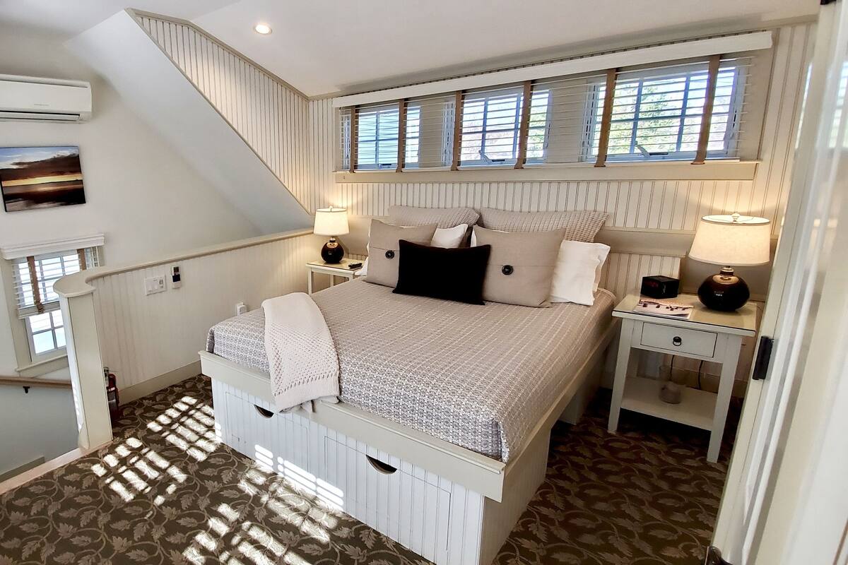 A bedroom with a large bed, two bedside tables with lamps, a window with shutters, a painting on the wall, and a staircase leading to another room.