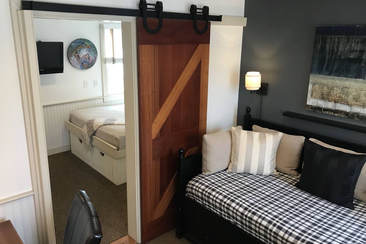 A bedroom with a bed, a nightstand, a wall-mounted lamp, and a door leading to another room.