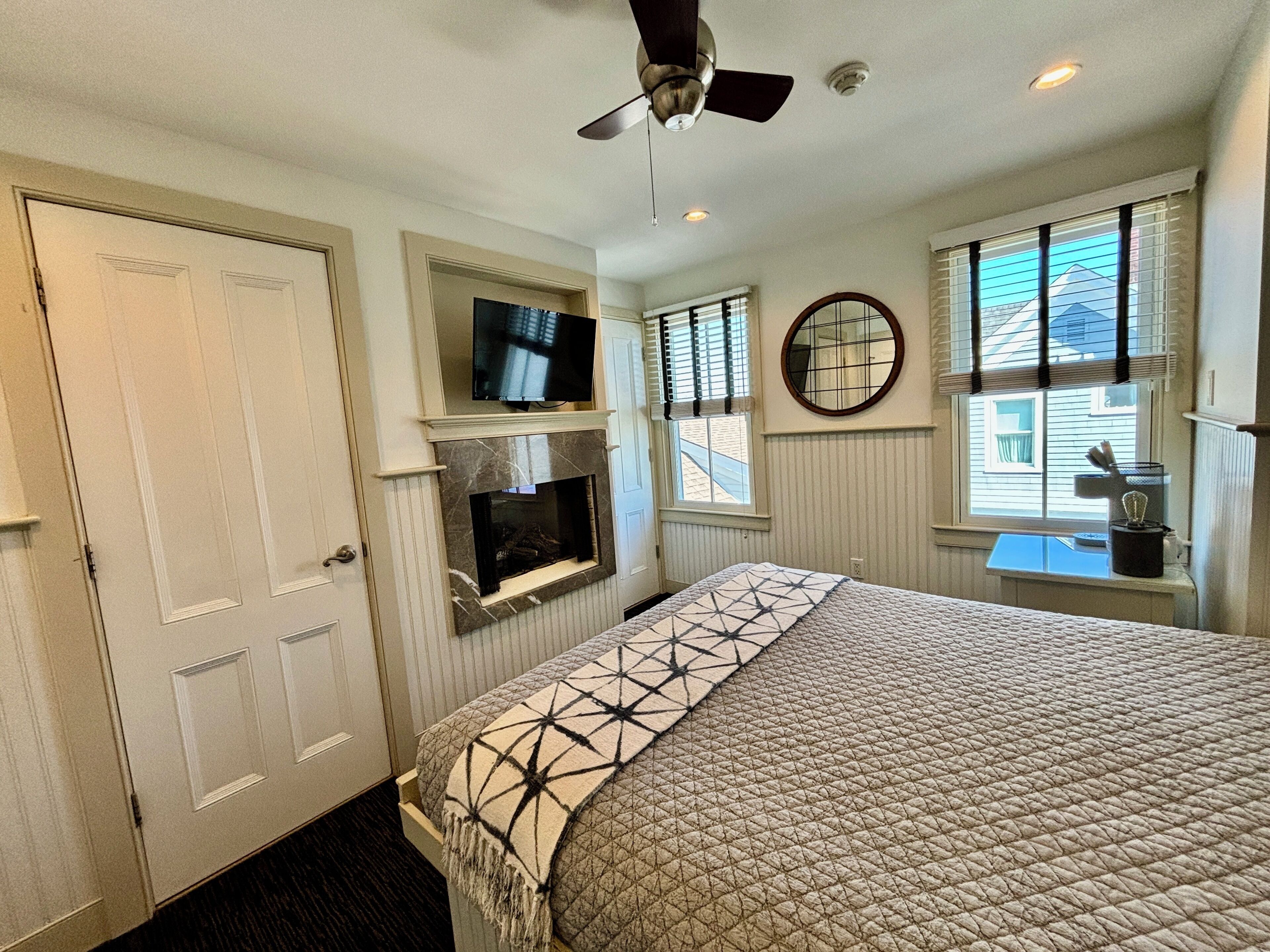 A bedroom with a bed, a television on a shelf, a fireplace, a ceiling fan, and a window with blinds.