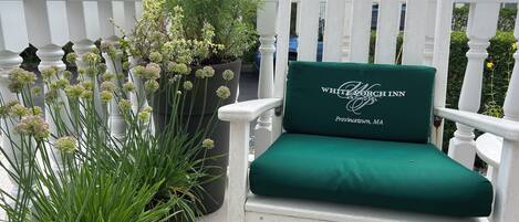 En vit stol med en grön kudde, där det står "WHITE PORCH INN, Provincetown, MA", placerad på ett trädäck med krukväxter.