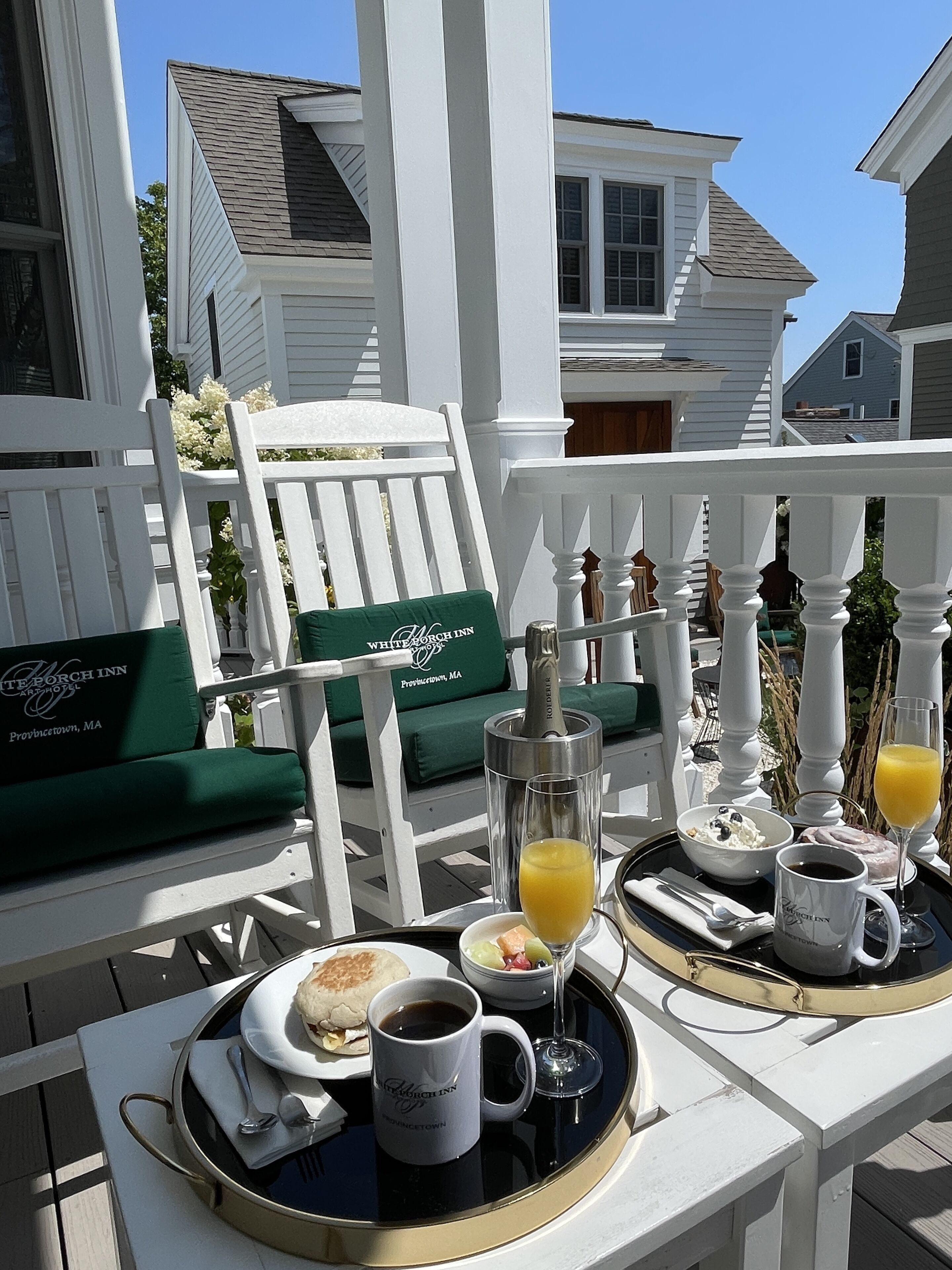 A breakfast tray on a white porch with a green cushion, a bottle, a glass of orange juice, a cup of coffee, a mug, and a plate with food.