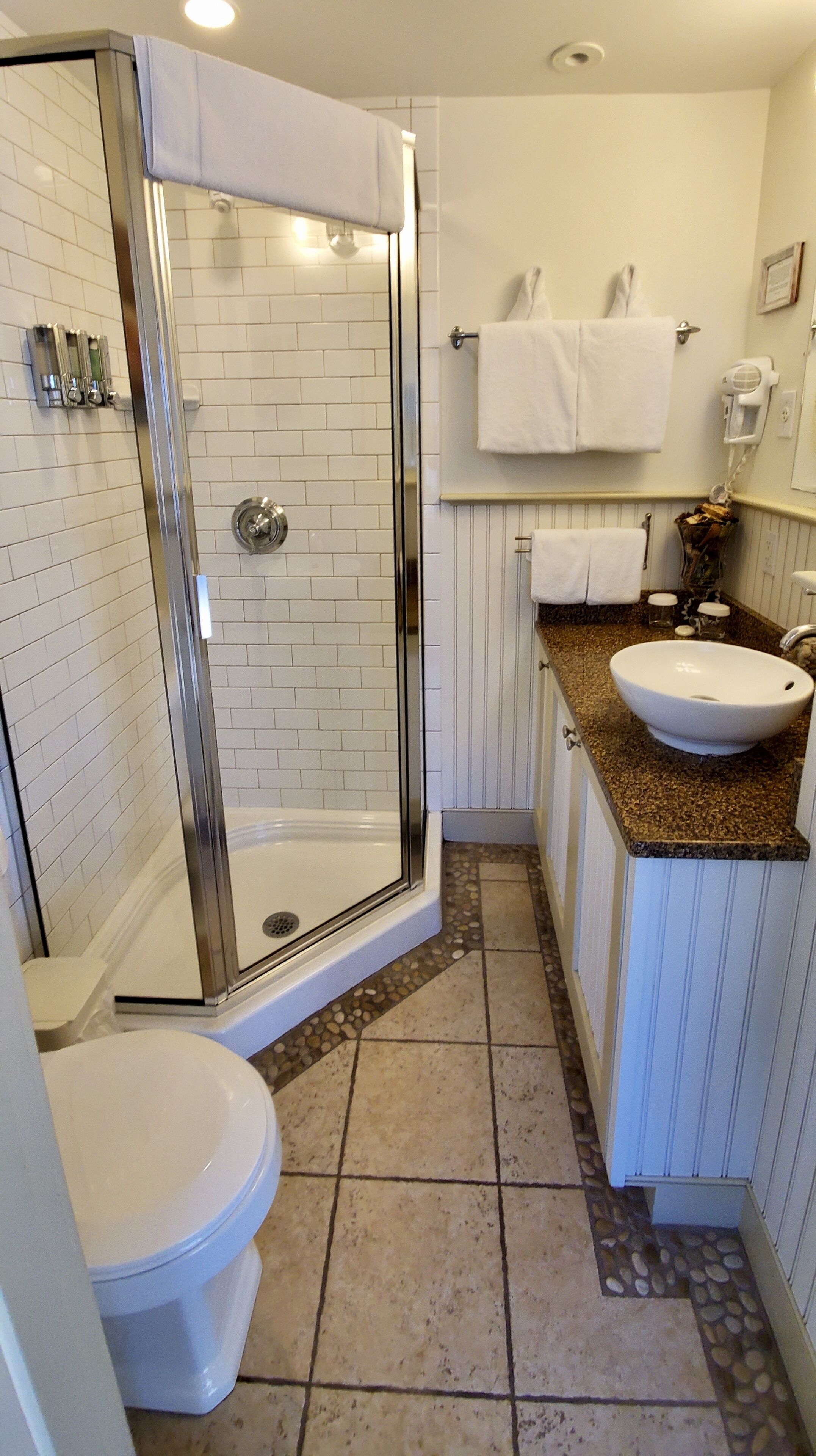 Un baño con ducha de vidrio, inodoro blanco, lavamanos con encimera de granito y toallero con varias toallas.