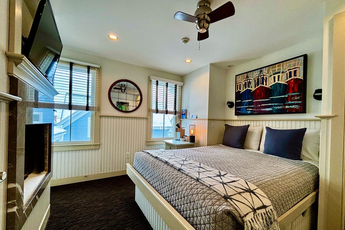 A bedroom with a bed, a ceiling fan, a TV, a painting, and a window.