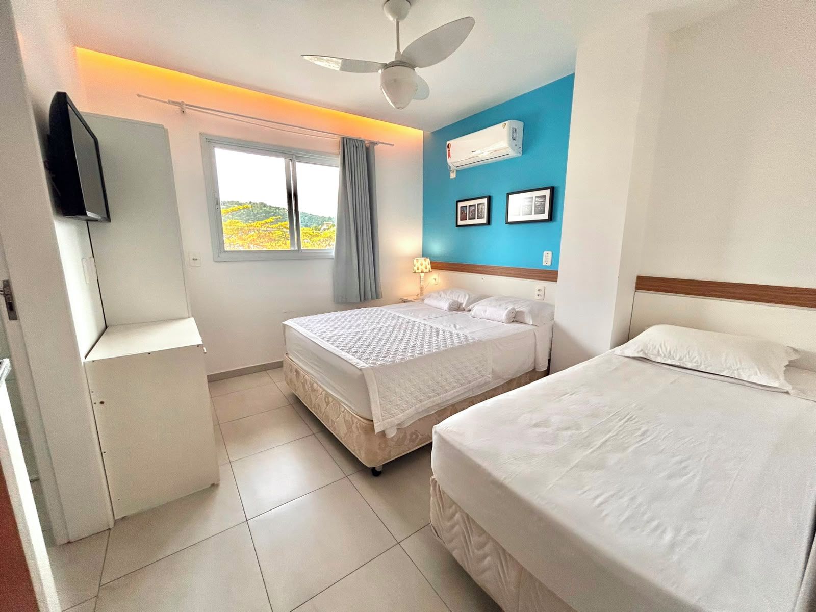 Habitación de hotel con dos camas, ventilador de techo, aire acondicionado, televisión y ventana con cortinas.