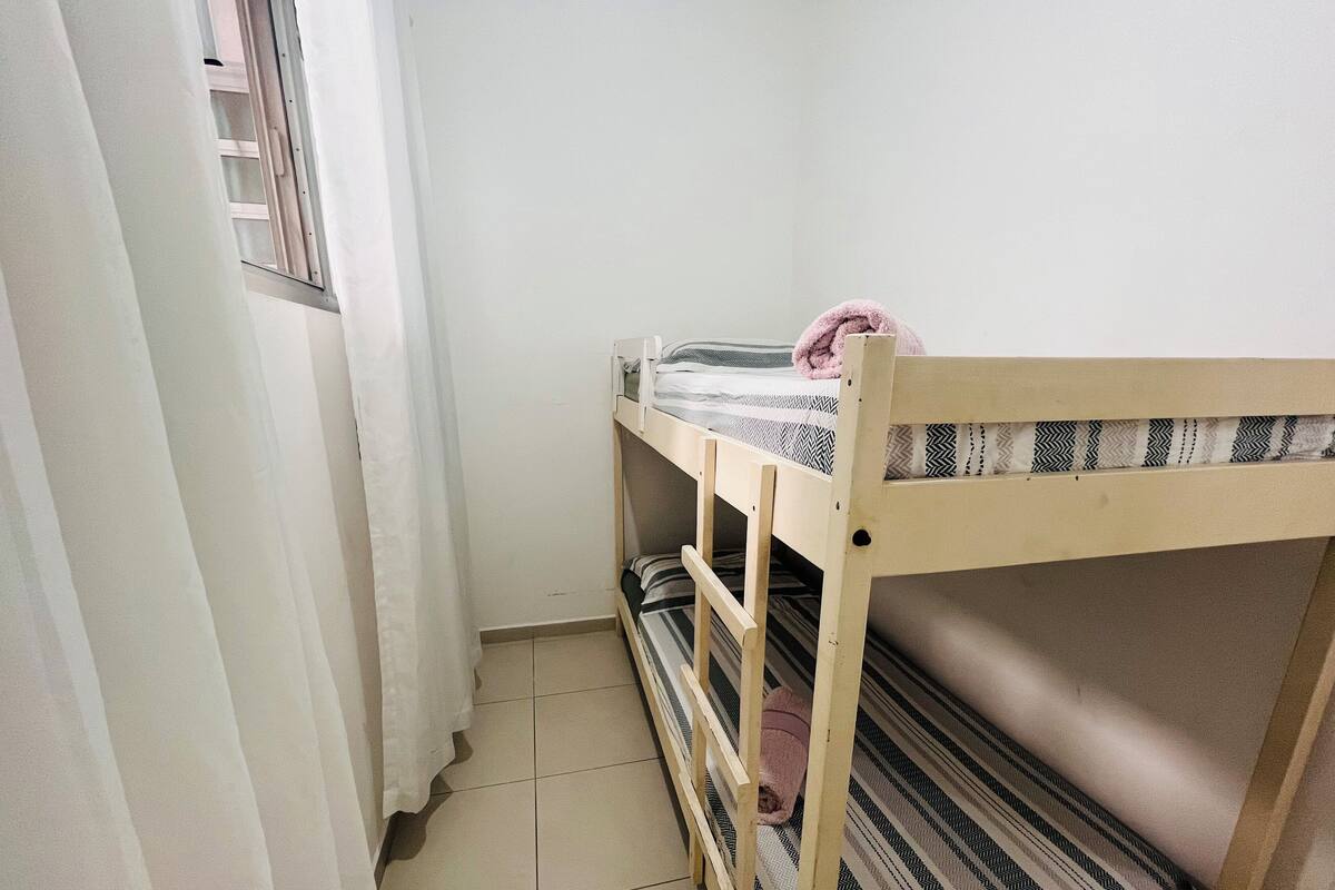Una cama individual superpuesta con un colchón a rayas, una ventana con cortina y un suelo de baldosas.