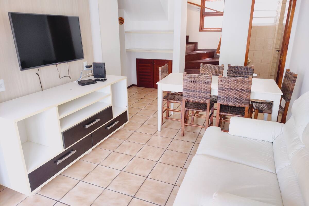 Una sala de estar con un sofá blanco, un televisor, una mesa de comedor con sillas de mimbre y una escalera que sube a un nivel superior.