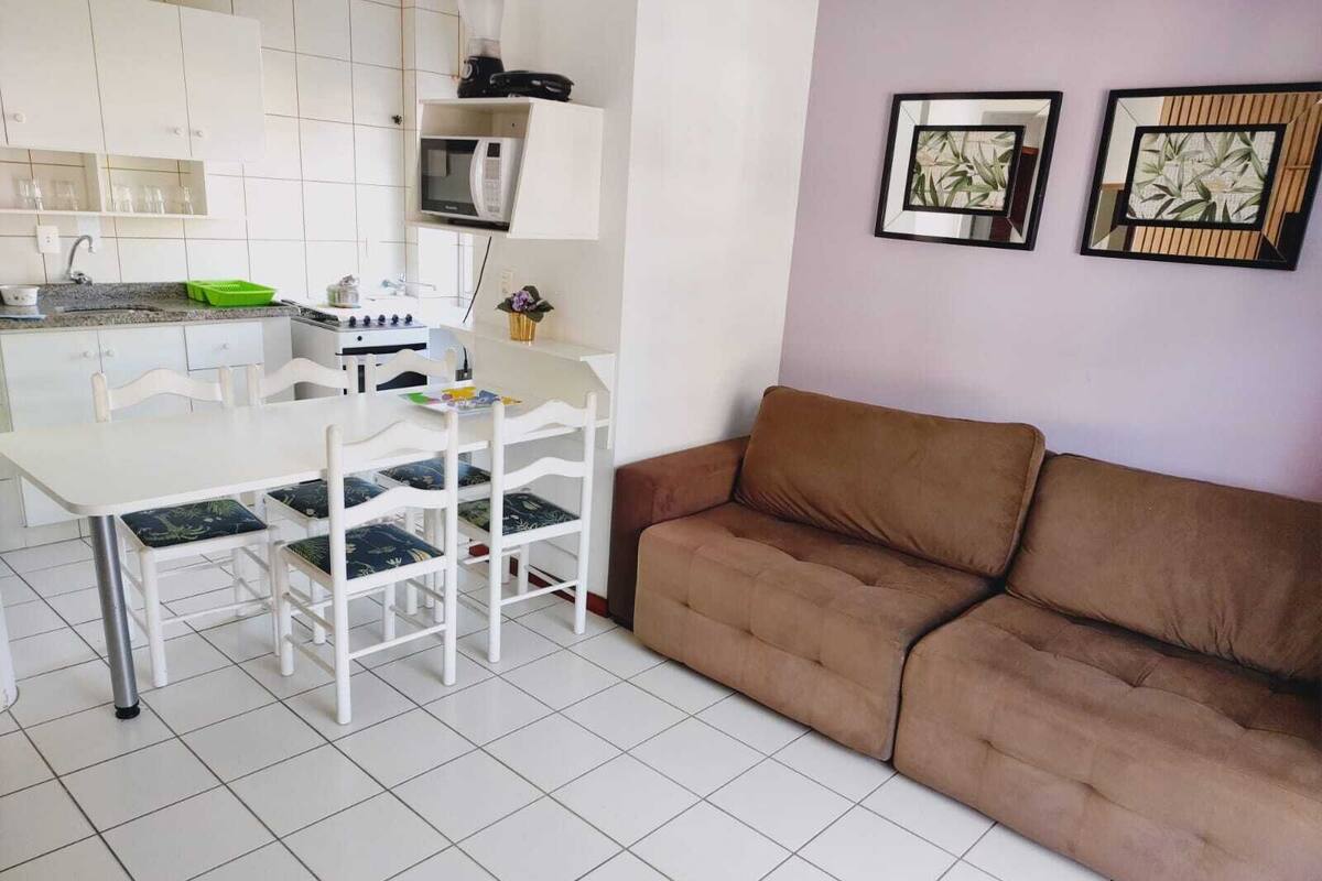 Un espacio habitable compacto con kitchenette, una zona de comedor con sillas blancas, un sofá marrón y dos obras de arte enmarcadas en la pared.
