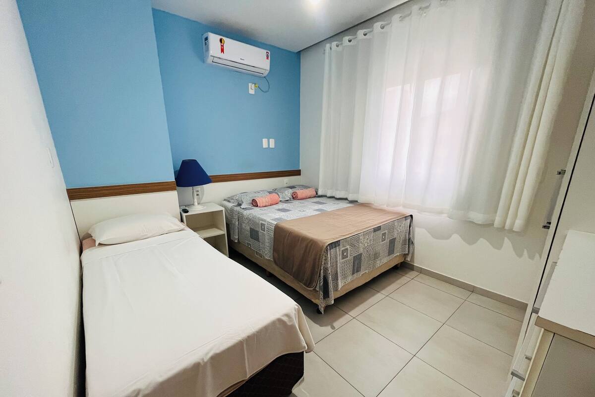 Habitación de hotel con dos camas, una pared azul, aire acondicionado, una mesita de noche con lámpara y una ventana con cortinas blancas.