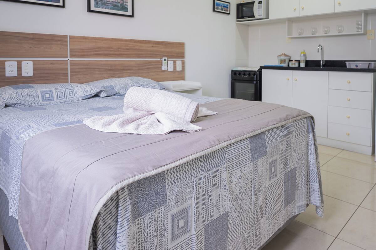 Un dormitorio con cama, mesita de noche, microondas y una zona de cocina con armarios y lavabo.
