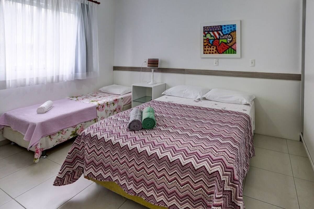 Habitación de hotel con dos camas, una mesita de noche con lámpara, un cuadro en la pared y una ventana con cortinas translúcidas.
