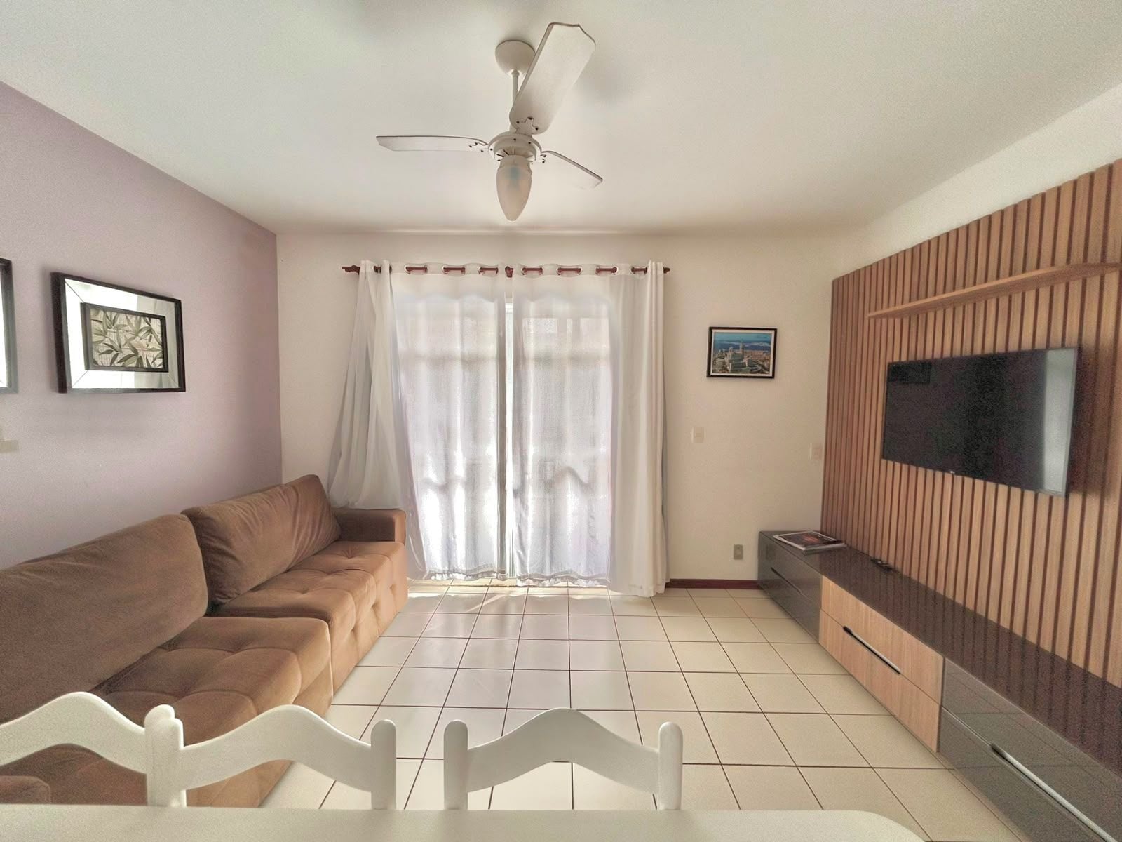 Ein Wohnzimmer mit einer Couch, einem an einer Holzwand befestigten Fernseher, einem Deckenventilator und einem Fenster mit Vorhängen.
