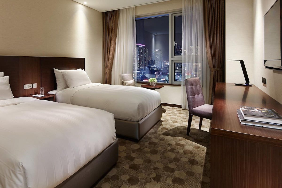 Quarto de hotel com duas camas, uma escrivaninha, uma cadeira e vista para a paisagem urbana à noite.