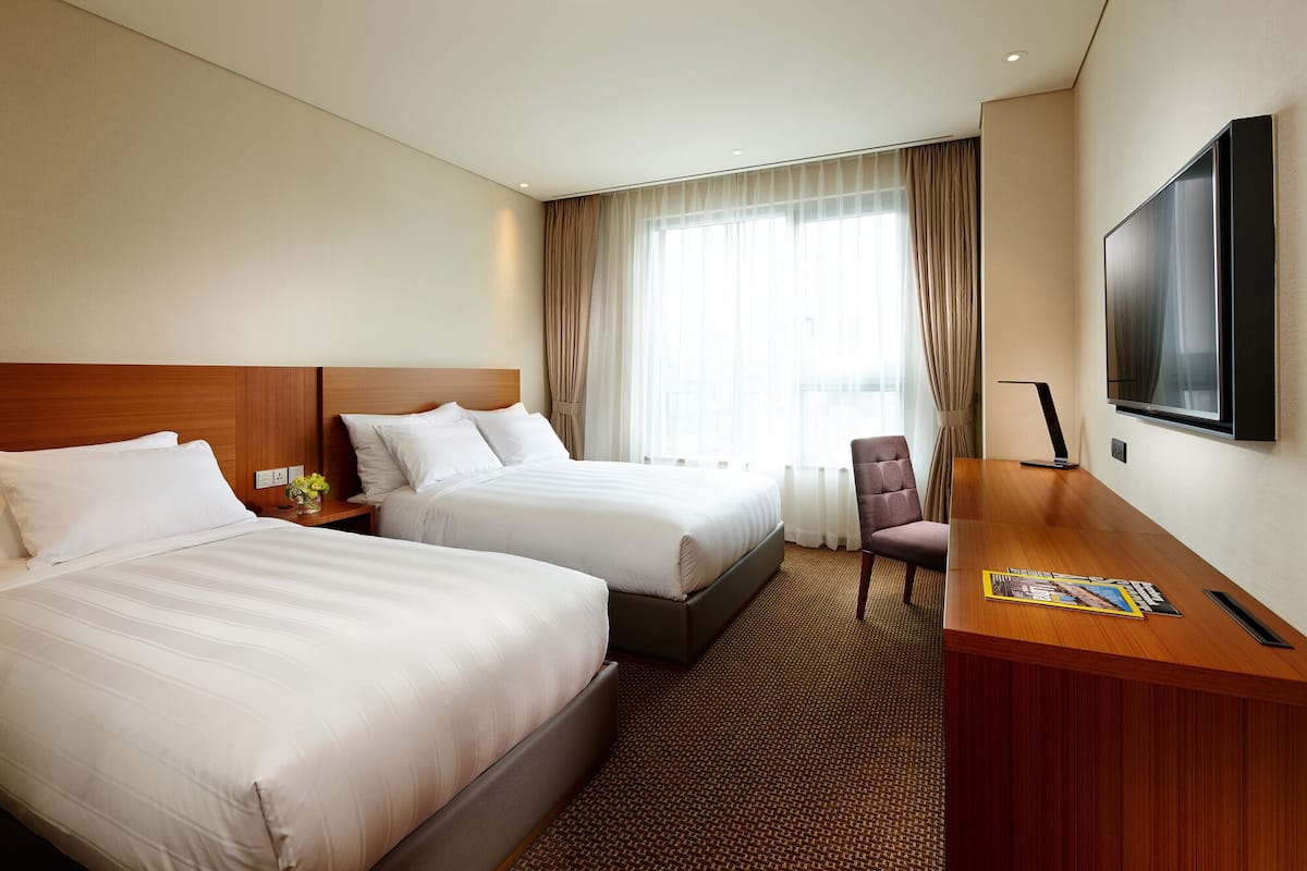 Quarto de hotel com duas camas, uma mesa, uma cadeira, televisão e janela com cortinas.