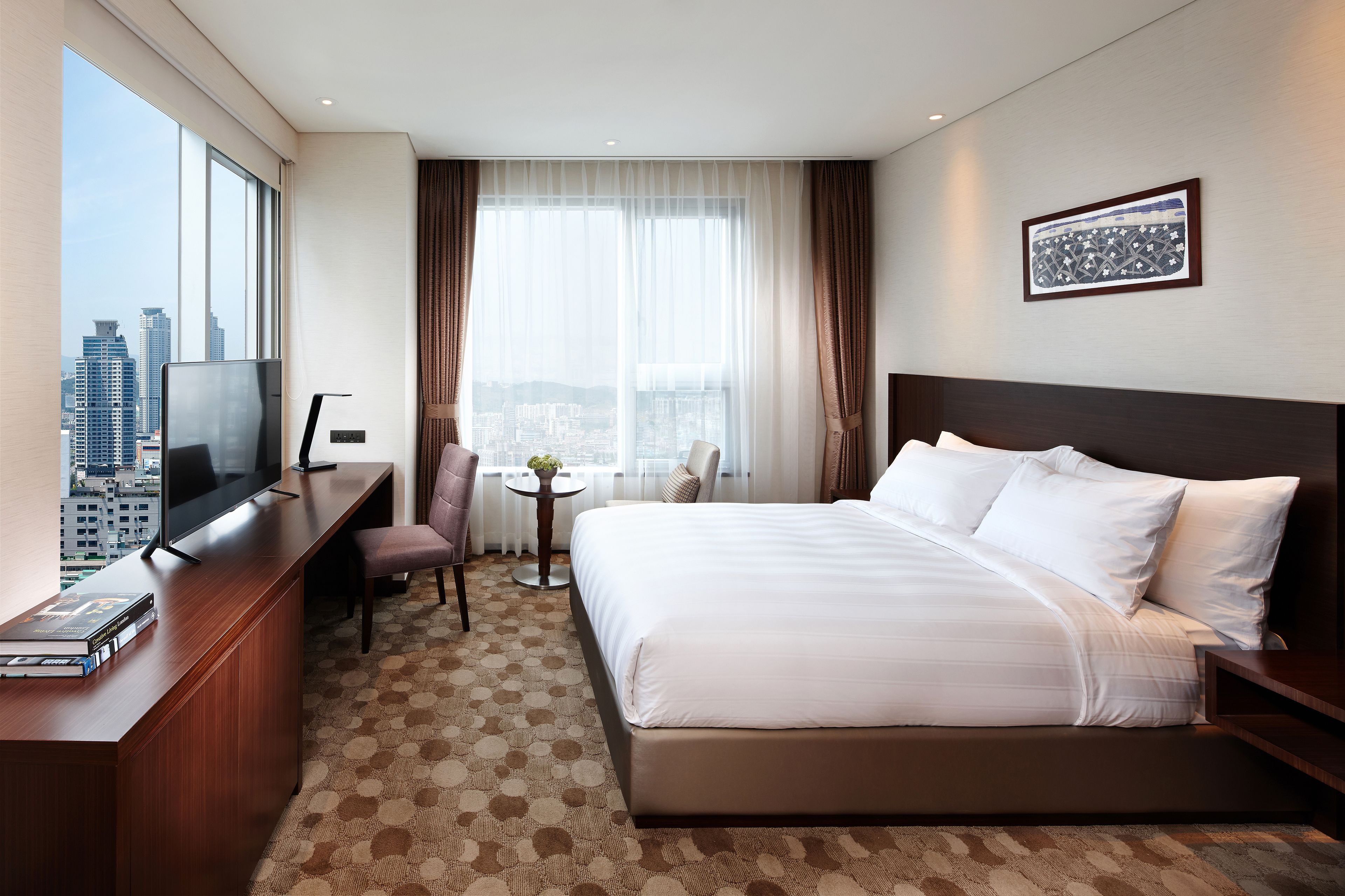 A hotel room with a large bed, a desk with a TV, a small table with a plant, and a city view.