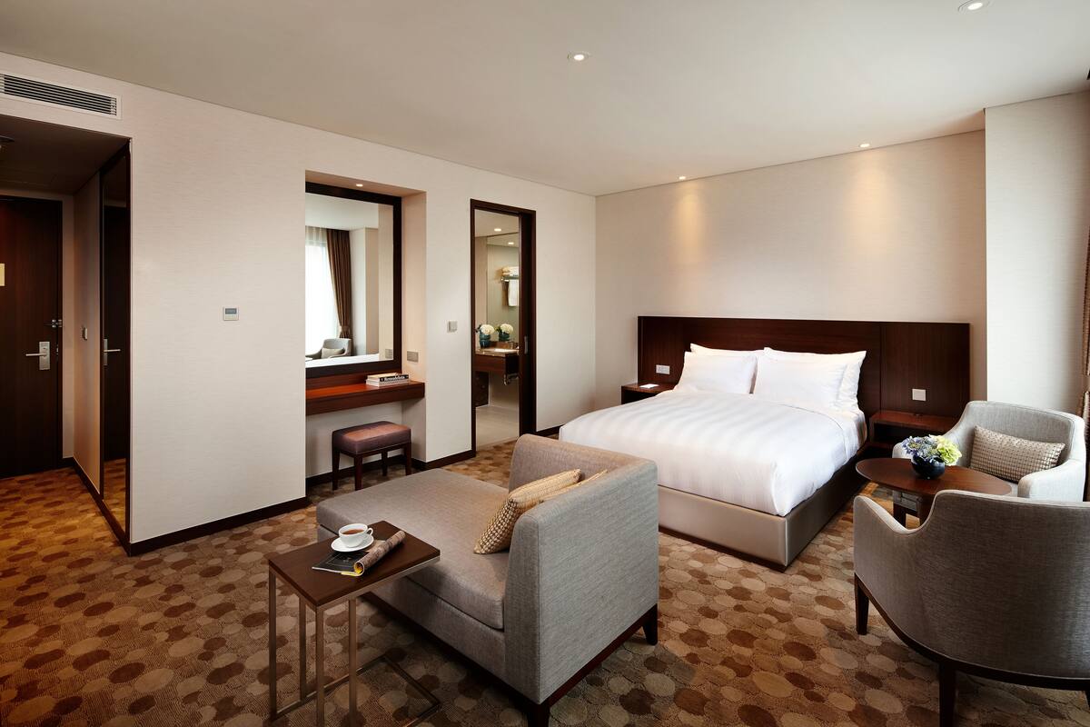 Quarto de hotel com uma cama grande, duas poltronas, uma mesa pequena e uma escrivaninha com cadeira.