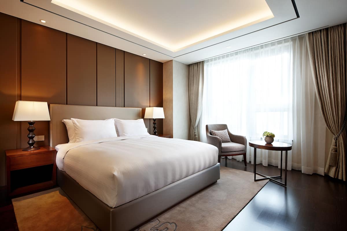 Quarto de hotel moderno com uma cama grande, abajures, uma mesa redonda com uma planta e vista para o exterior através de cortinas leves.