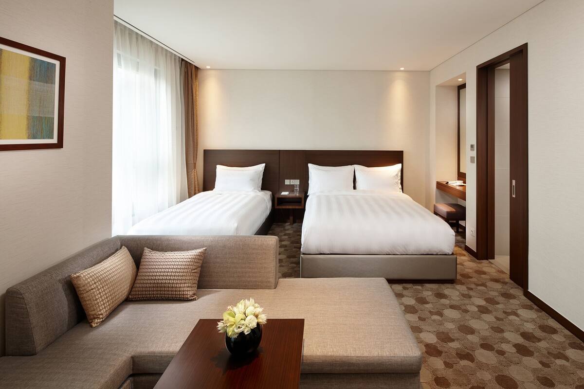 Quarto de hotel com duas camas, um sofá, uma mesa de centro e uma janela grande com cortinas.