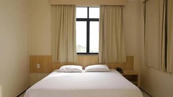 Quarto de hotel com cama, mesa de cabeceira, janela com cortinas e cabeceira de madeira.