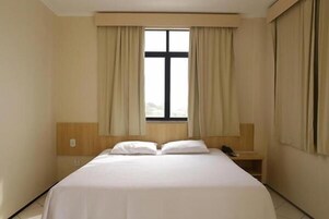 Quarto de hotel com cama, mesa de cabeceira, janela com cortinas e cabeceira de madeira.