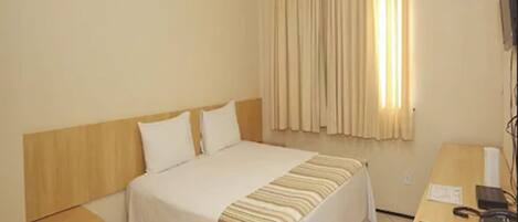 A hotel room with a bed, a nightstand, a desk with a telephone, and a window with curtains.