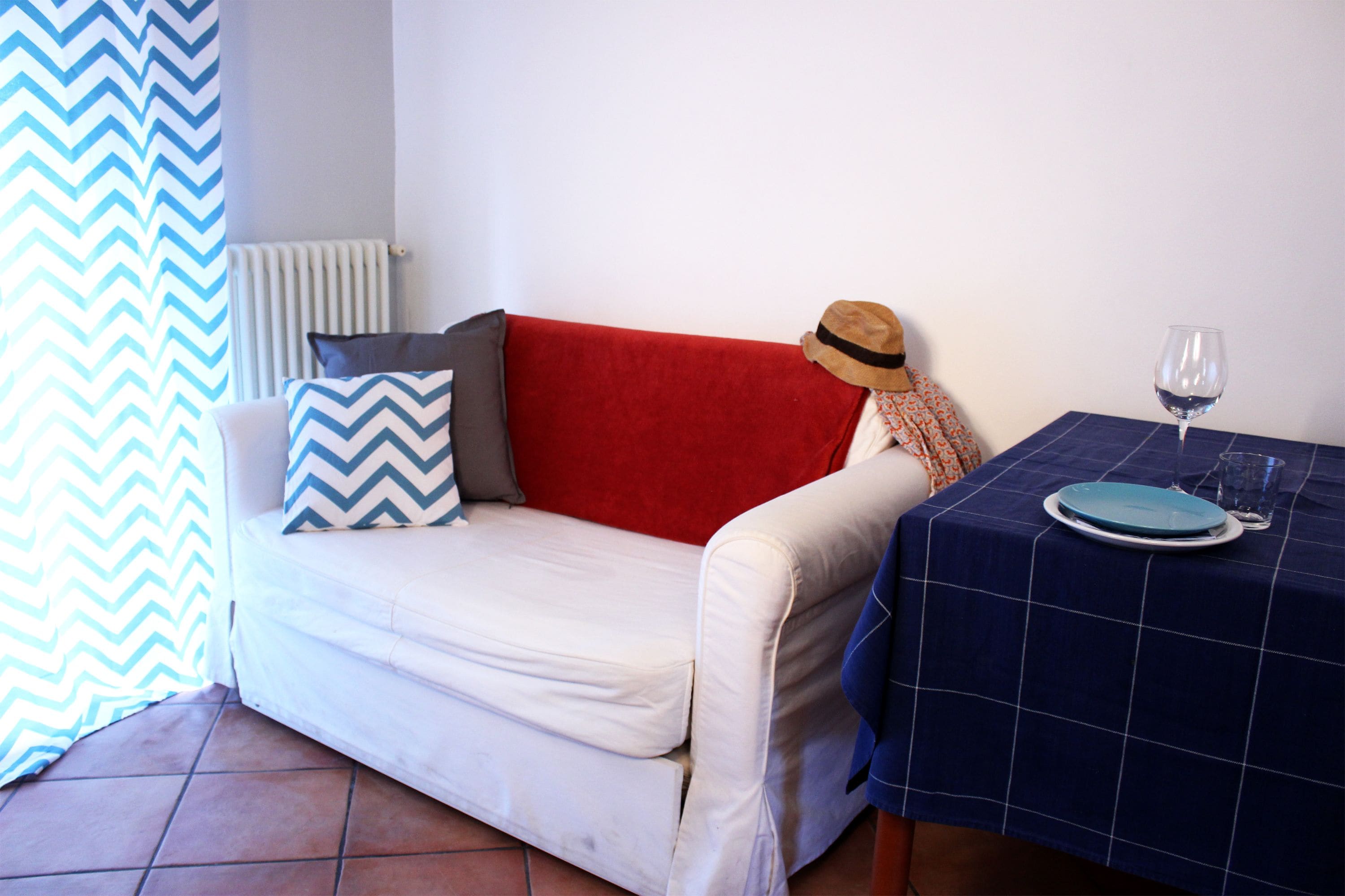 A room with a white sofa, a red cushion, a blue and white chevron pillow, a hat on the sofa, a table with a blue tablecloth, a plate, and a wine glass.