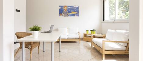 A modern, well-lit room with a white table, a laptop, a potted plant, a sofa, and a bed.